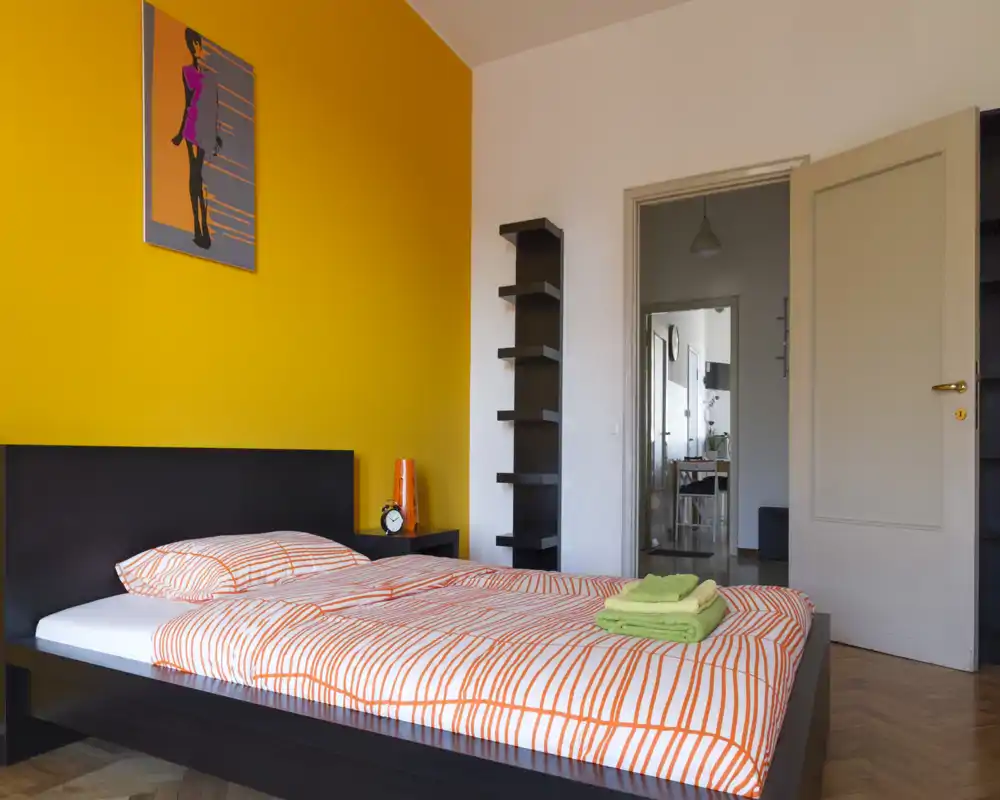 Camera da letto moderna e luminosa con letto in legno scuro basso, biancheria arancione e una parete gialla vivace. L'ambiente è ordinato e accogliente con luce naturale.
