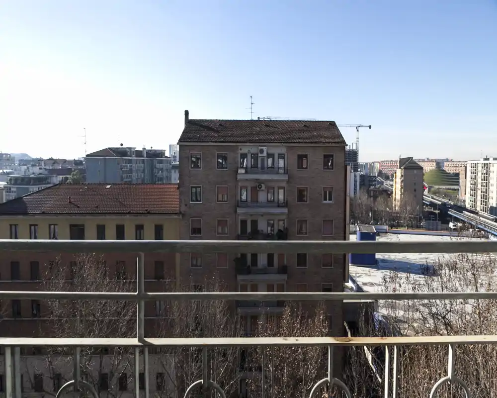 Vista dal balcone con ringhiera in metallo in primo piano e palazzi residenziali di fronte, cielo limpido.