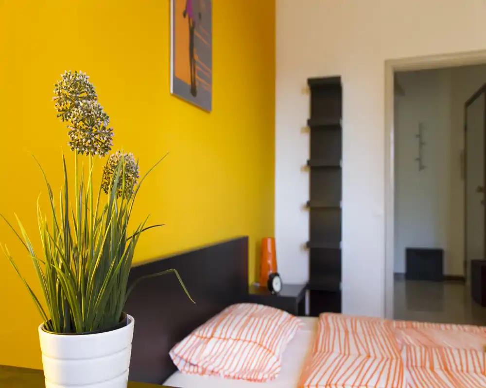 Camera da letto luminosa e moderna con parete gialla, letto in ordine con biancheria a righe e accessori sul comodino, oltre a una pianta decorativa in primo piano.