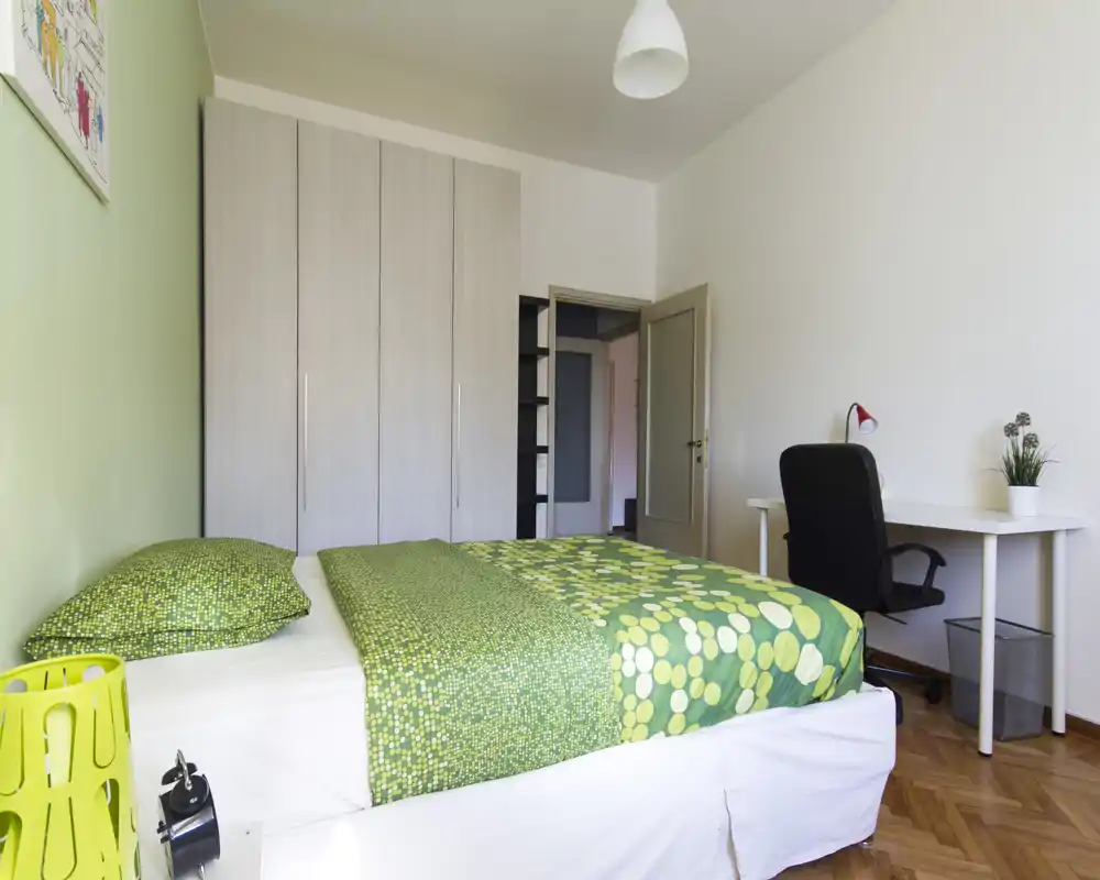 Dormitorio luminoso y ordenado con cama doble con ropa de cama verde estampada, un gran armario y un escritorio sencillo con silla — ideal para estancias confortables y teletrabajo.