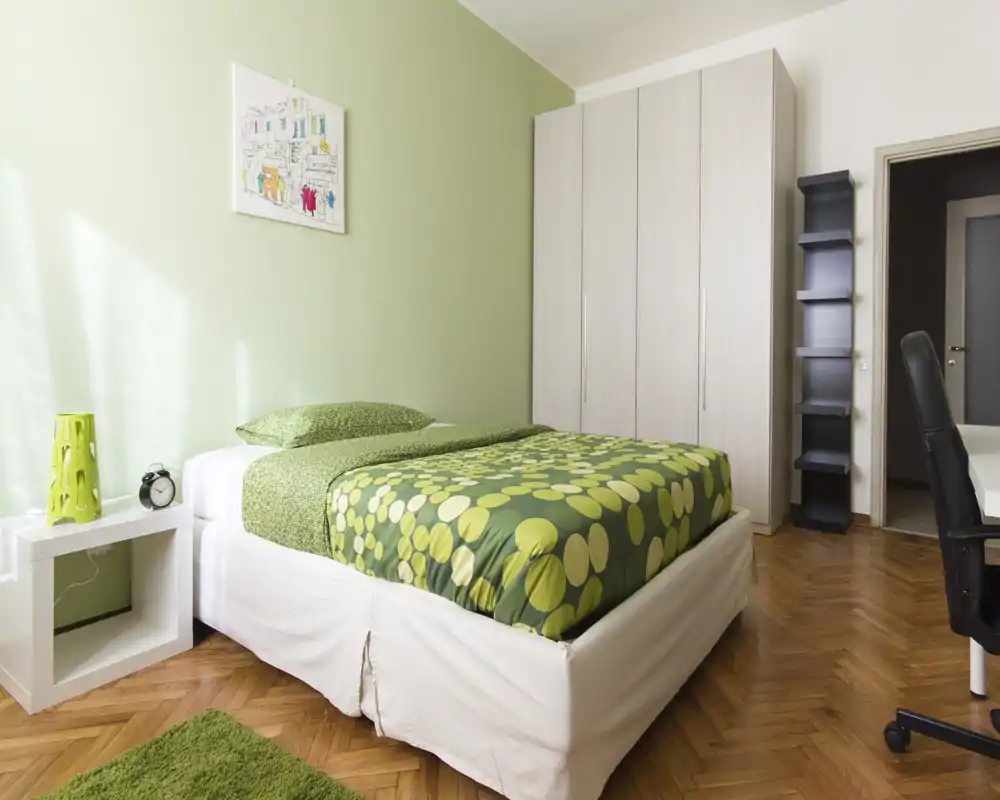 Dormitorio luminoso y ordenado con cama doble vestida con ropa de cama verde estampada, armario, zona de escritorio y suelo de parquet — ideal para estancias cómodas.