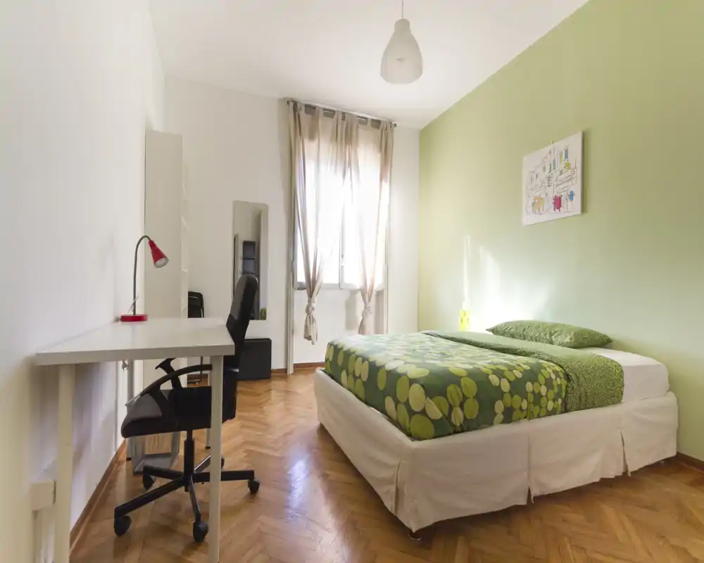 Habitación luminosa y bien decorada con cama doble con ropa de cama verde estampada, escritorio y silla junto a la pared y luz natural de una ventana grande.