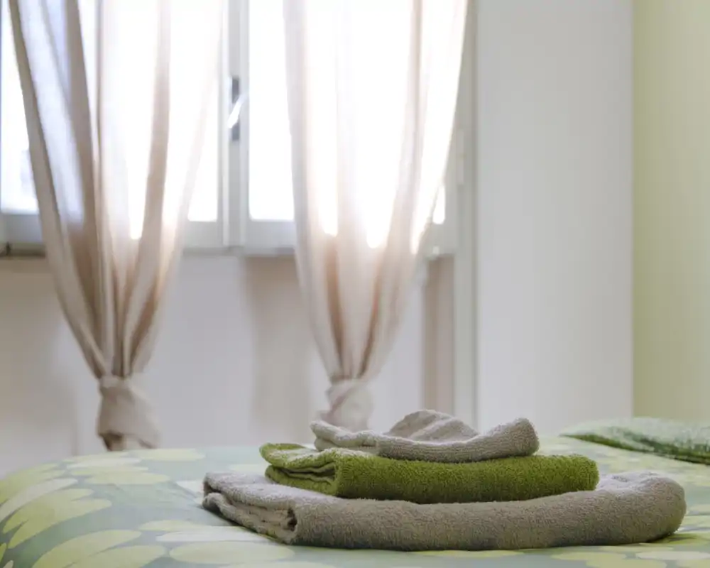 Dormitorio luminoso y bien presentado con la cama hecha, toallas dobladas y cortinas que dejan pasar la luz natural: acogedor.