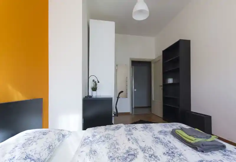 Camera da letto luminosa e ordinata con letto matrimoniale in primo piano, parete arancione a contrasto e mobili neri minimal. Ambiente ben illuminato e arredamento essenziale, adatto per annunci in affitto.