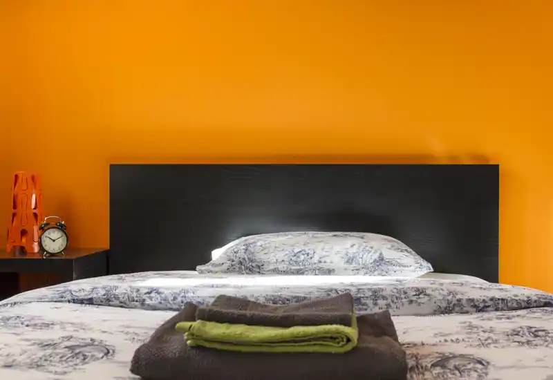 Camera da letto luminosa e ben arredata con un grande letto centrato contro una testiera scura e una parete arancione vivace. Lenzuola nitide, asciugamani piegati sul letto e una sveglia sul comodino creano un'atmosfera accogliente e moderna.