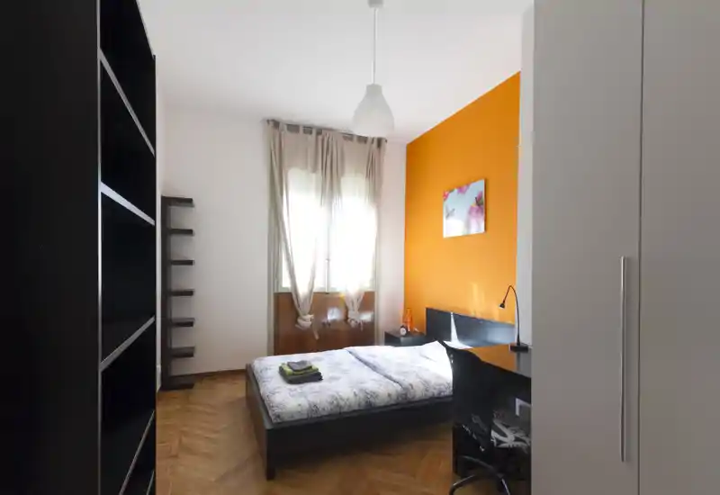 Camera da letto moderna e luminosa con letto matrimoniale, comodino e scrivania; parete arancione e luce naturale dalla finestra rendono l'ambiente accogliente.