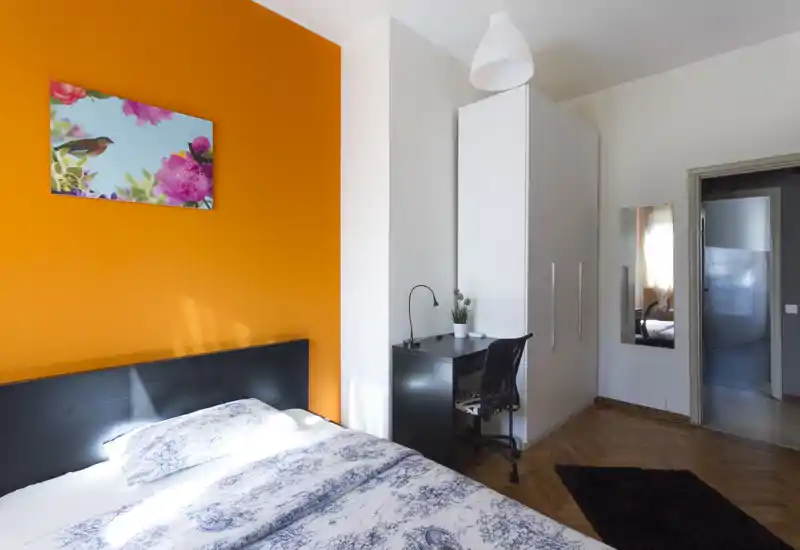 Camera da letto luminosa e moderna con parete arancione a contrasto, letto matrimoniale con biancheria a fantasia, armadio e scrivania: ideale per soggiorni brevi e lavoro in stanza.