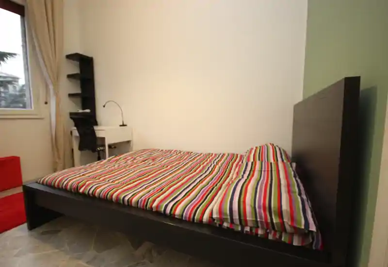 Dormitorio moderno y luminoso con una cama grande de madera oscura y ropa de cama a rayas coloridas; un pequeño escritorio junto a la ventana proporciona espacio de trabajo con luz natural.