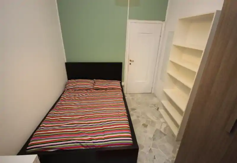 Dormitorio compacto con cama colocada junto a una pared verde menta, ropa de cama a rayas, estantería blanca y suelo efecto mármol — luminoso y de mobiliario sencillo.