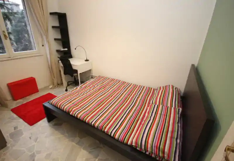 Dormitorio luminoso y ordenado con cama de madera oscura, edredón de rayas coloridas, pequeño escritorio y ventana con luz natural — ideal para un alquiler urbano acogedor.