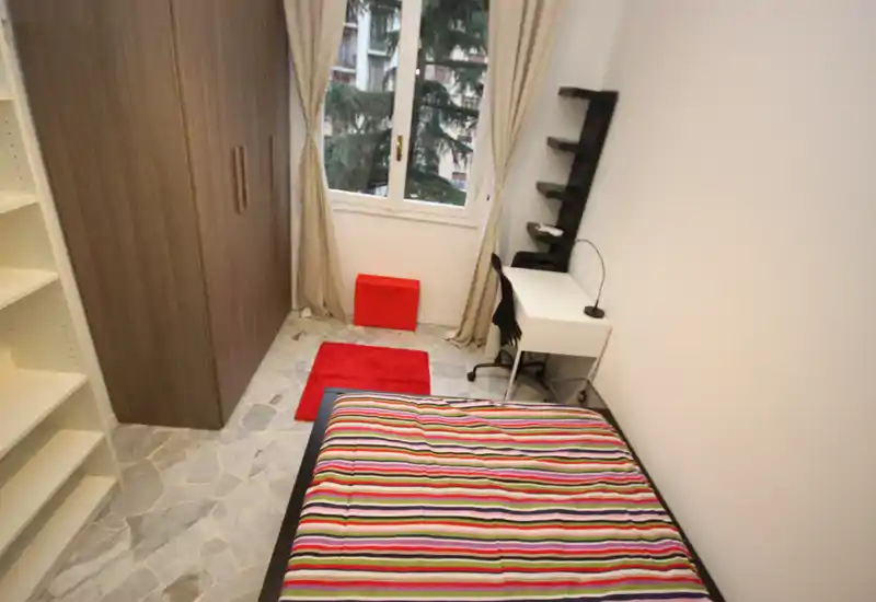 Dormitorio individual luminoso y ordenado con cama de rayas coloridas, armario empotrado, escritorio y luz natural de una ventana grande.