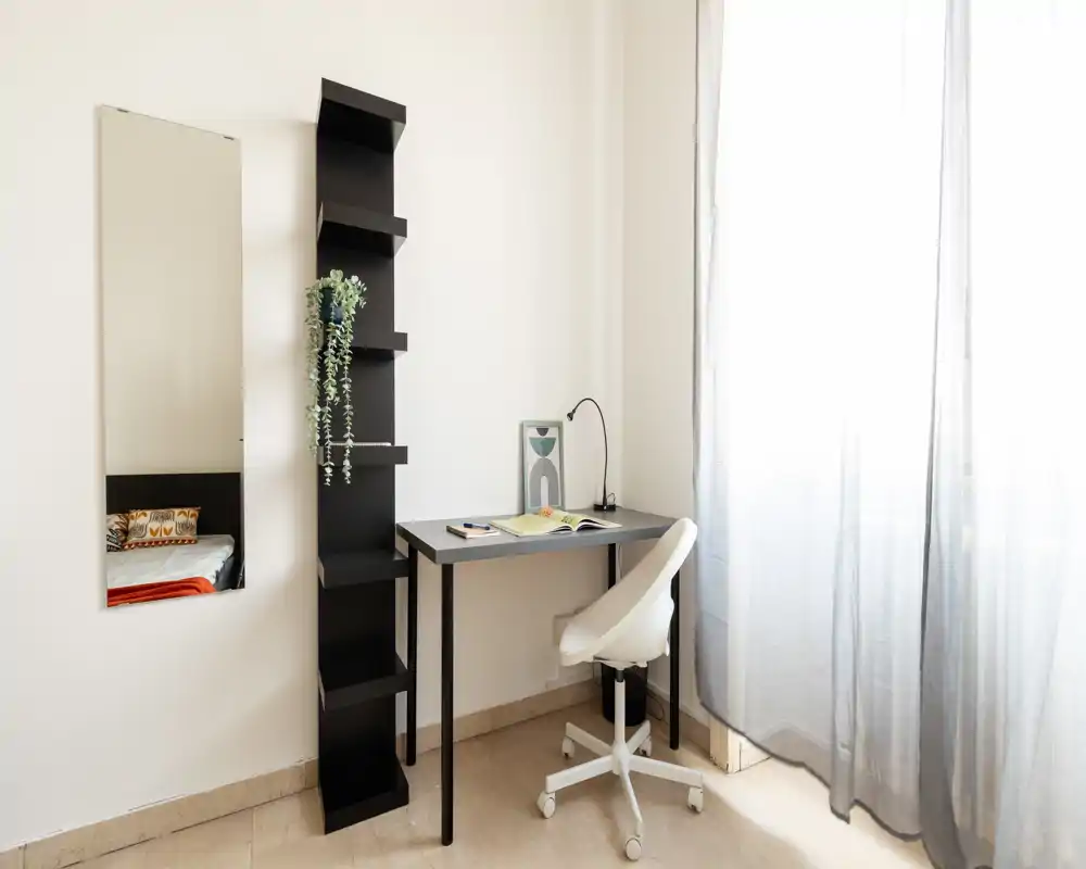 Rincón de dormitorio luminoso y minimalista con escritorio compacto y silla giratoria junto a una gran ventana; un espejo a la izquierda refleja parte de la cama y cojines decorativos.
