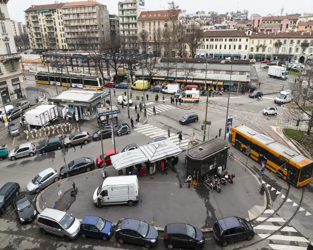 Scena esterna a livello strada che mostra un angolo urbano trafficato con auto parcheggiate, una bancarella con tendoni, autobus e una fermata del tram sullo sfondo.
