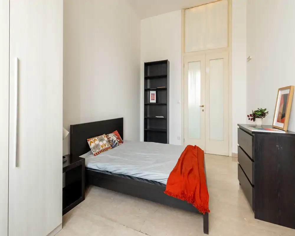 Dormitorio luminoso y minimalista con cama doble, cojines decorativos y una cálida manta roja, muebles de almacenamiento y luz natural.