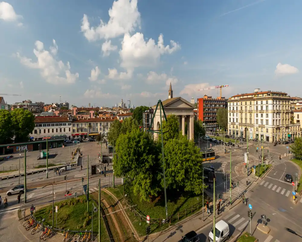 Vista panorámica de una plaza y calles de la ciudad con vías de tranvía, árboles frondosos y edificios clásicos bajo un cielo despejado — ideal para destacar la ubicación y el transporte cercano.
