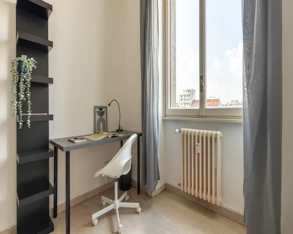 Rincón de dormitorio compacto y luminoso con un pequeño escritorio y silla junto a la ventana que deja entrar luz natural; mobiliario moderno y minimalista y radiador bajo la ventana.
