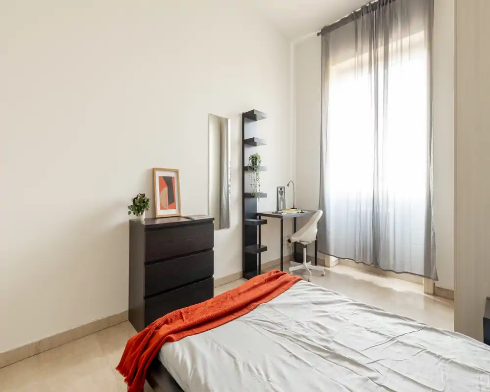 Dormitorio luminoso y minimalista con cama visible, ropa de cama clara y una manta naranja; gran ventana que proporciona luz natural y un pequeño escritorio junto a la ventana.