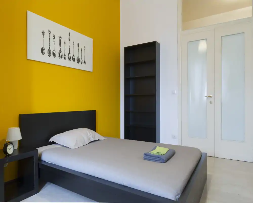 Camera da letto moderna e minimal con letto completo, parete d'accento gialla, comodino e libreria — luminosa, pulita e pronta per gli ospiti.