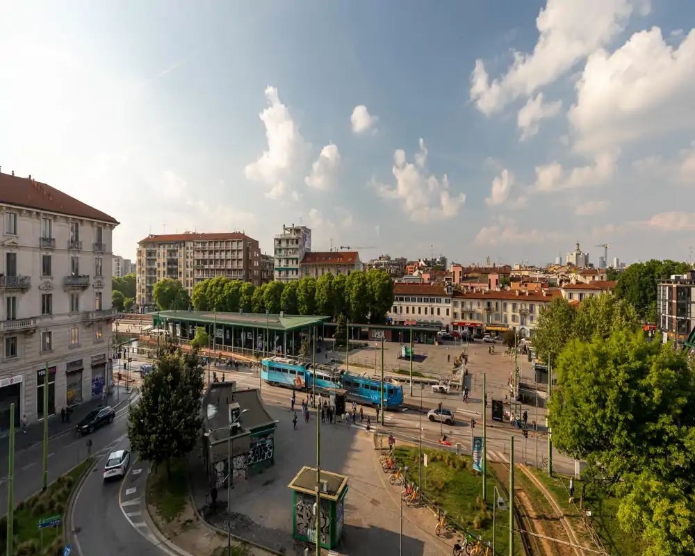 Vista urbana soleada de una parada de tranvía y la plaza circundante con edificios residenciales y árboles, ideal para destacar las conexiones de transporte público.
