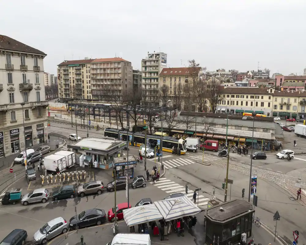 Vivace scena urbana con tram, auto e un piccolo mercato all'aperto; ottima panoramica sui trasporti pubblici e i negozi locali.