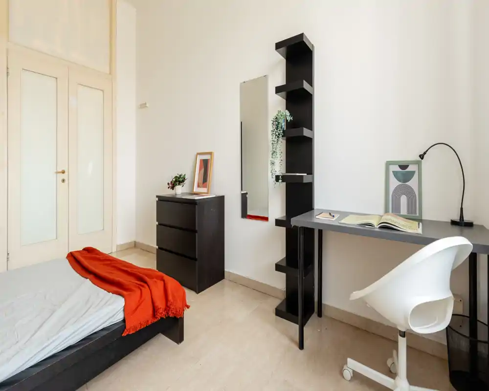 Dormitorio luminoso y minimalista con cama baja, cómoda negra y una zona de trabajo ordenada con estanterías y silla — ideal para una persona o estudiante.