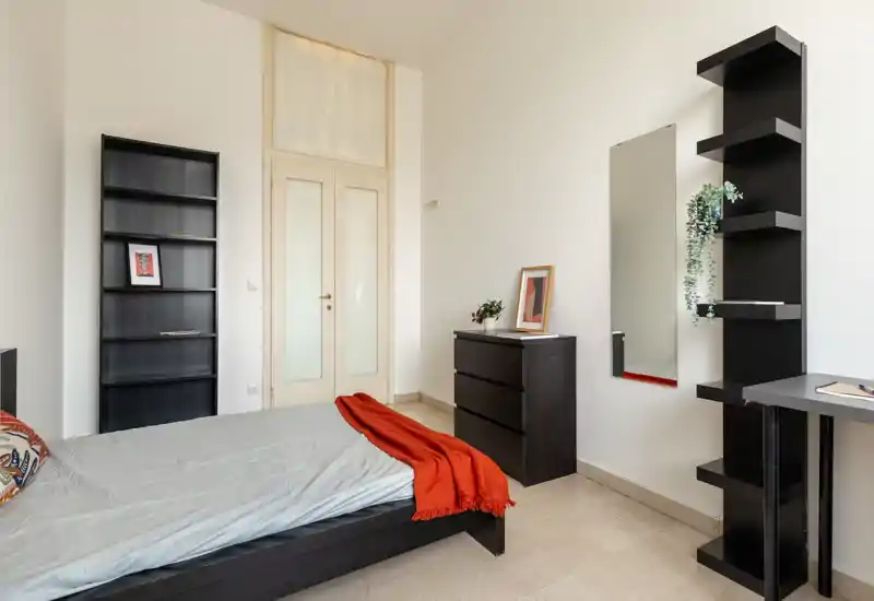 Camera da letto luminosa e minimal con letto basso, biancheria ordinata e un caldo plaid arancione; scaffalature semplici, comò e uno specchio a parete completano l'ambiente funzionale e arioso.