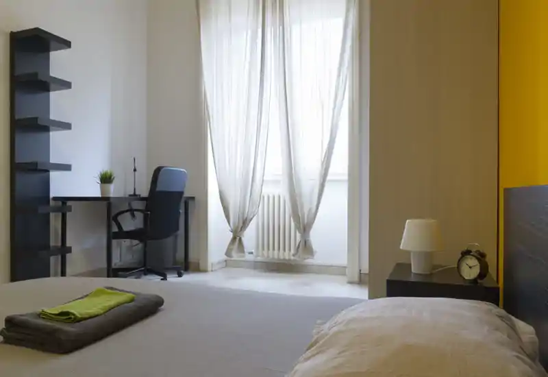 Camera da letto moderna e luminosa con letto ordinato in primo piano, comodino con lampada e sveglia, e una piccola scrivania vicino alla finestra che diffonde luce naturale soffusa.