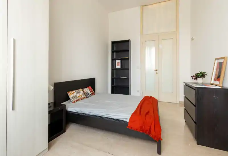 Bright, minimally styled bedroom with a double bed, decorative cushions and a warm red throw, plus storage furniture and natural light.