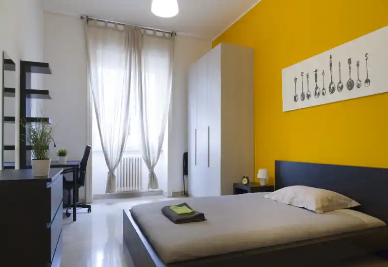 Camera da letto luminosa e moderna con letto matrimoniale, parete gialla accentuata, armadio e scrivania vicino a una finestra che illumina l'ambiente.