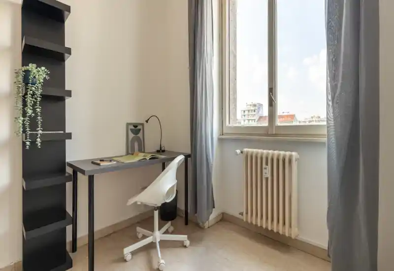 Compact, bright bedroom corner with a small desk and chair by a window letting in natural light; minimal, modern furnishings and a radiator under the window.