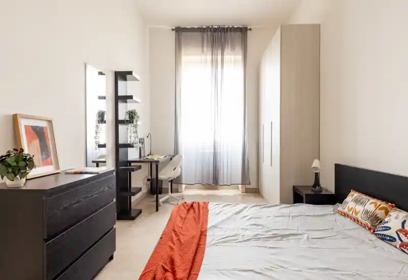 Camera da letto moderna e luminosa con letto ampio, armadio, comò e una scrivania vicino alla finestra. Toni neutri e dettagli curati (coperta e cuscini) creano un ambiente accogliente, perfetto per annunci.
