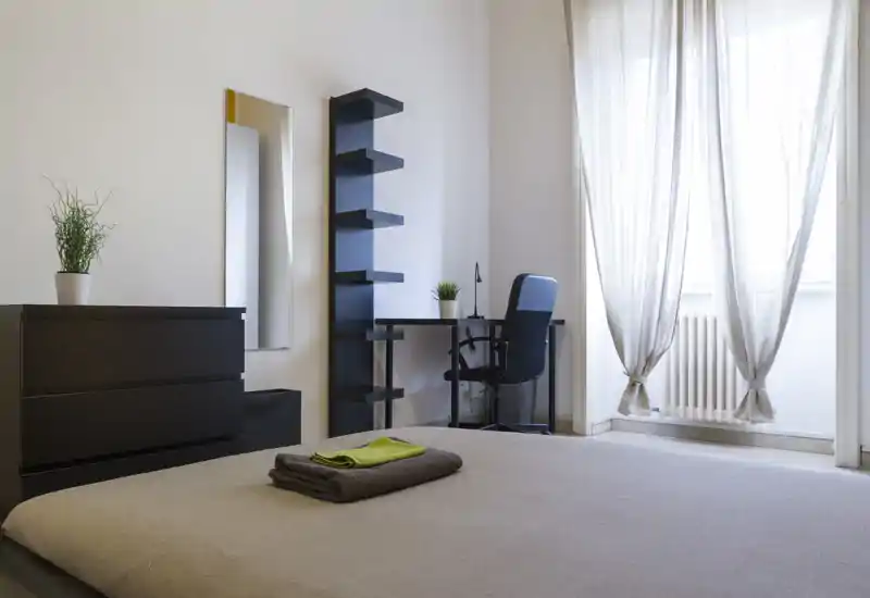 Camera da letto minimal e luminosa con letto ben rifatto in primo piano, scrivania e scaffalatura vicino alla finestra e luce naturale che crea un'atmosfera ariosa e moderna.