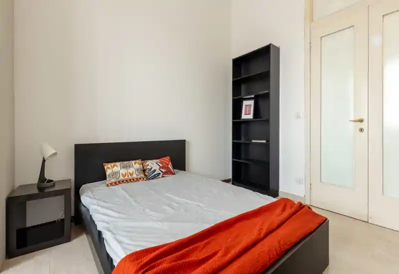 Camera da letto minimalista con letto matrimoniale, cuscini decorativi e un caldo plaid arancione; linee pulite, comodino con lampada e una alta libreria vicino alla porta.