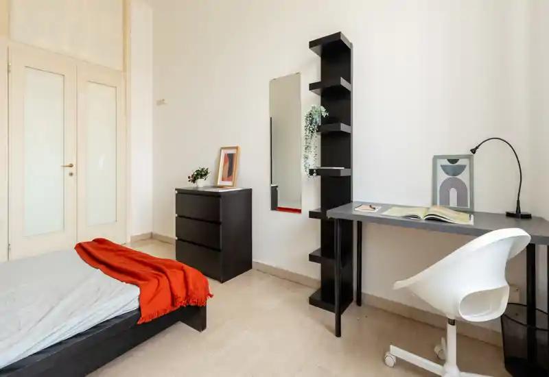 Camera da letto luminosa e minimale con letto basso, comò nero e una postazione di lavoro ordinata con mensole e sedia — ideale per single o studenti.