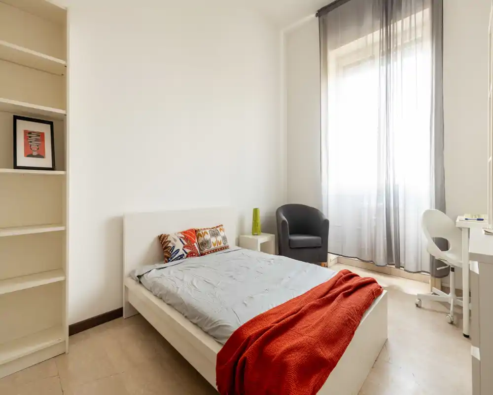 Dormitorio luminoso y ordenado con cama doble, cojines coloridos y una manta roja; un sillón cómodo y un pequeño escritorio junto a la ventana lo hacen funcional para descansar y trabajar.
