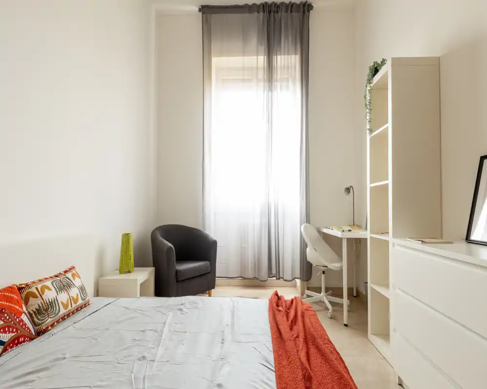 Dormitorio luminoso y minimalista con una cama cómoda, cojines decorativos y una cálida manta naranja, un sillón junto a la ventana y un escritorio compacto.