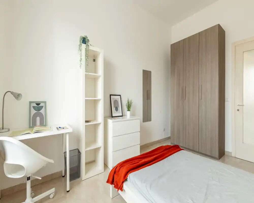 Dormitorio moderno y luminoso con cama ordenada y manta roja, armario alto, estantería y escritorio — líneas limpias y luz natural que lo hacen atractivo para un anuncio de alquiler.