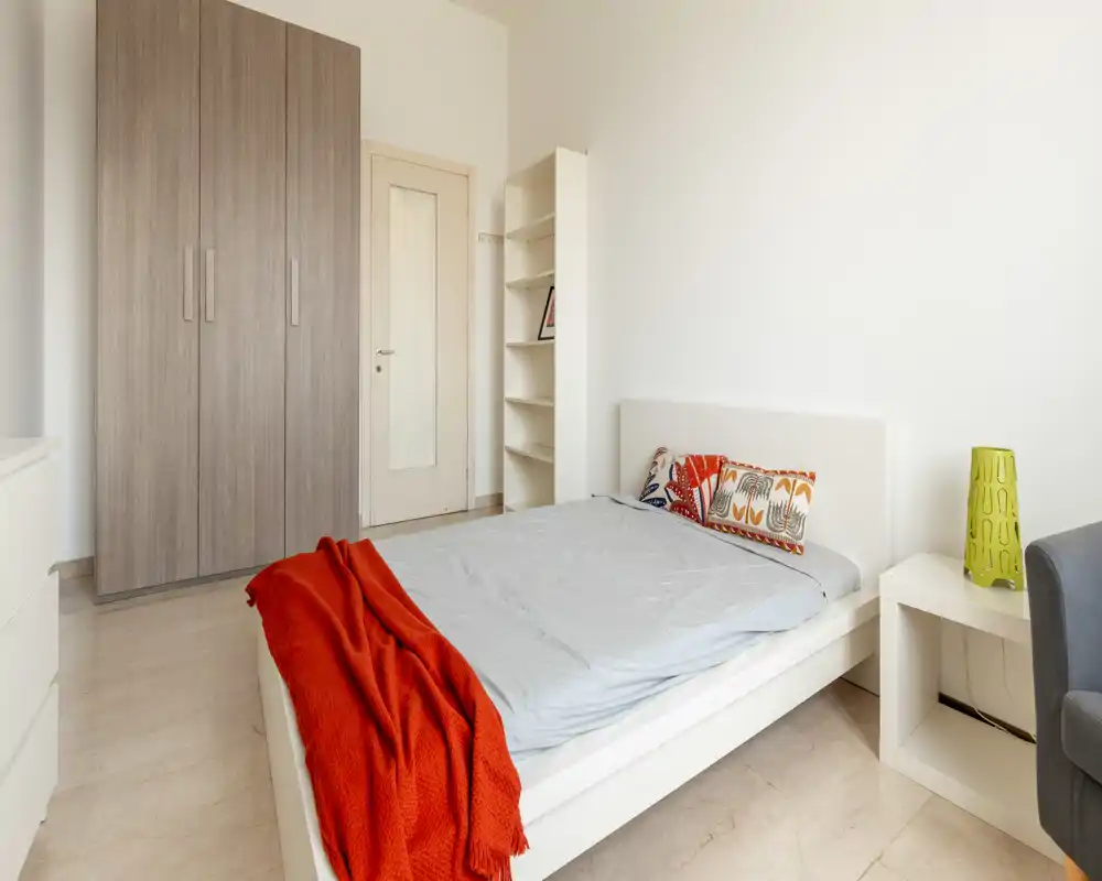 Dormitorio luminoso y minimalista con cama plataforma blanca, cojines coloridos y una manta roja. La habitación cuenta con un gran armario, mesita y estanterías sencillas — perfecta para un anuncio de alquiler moderno.