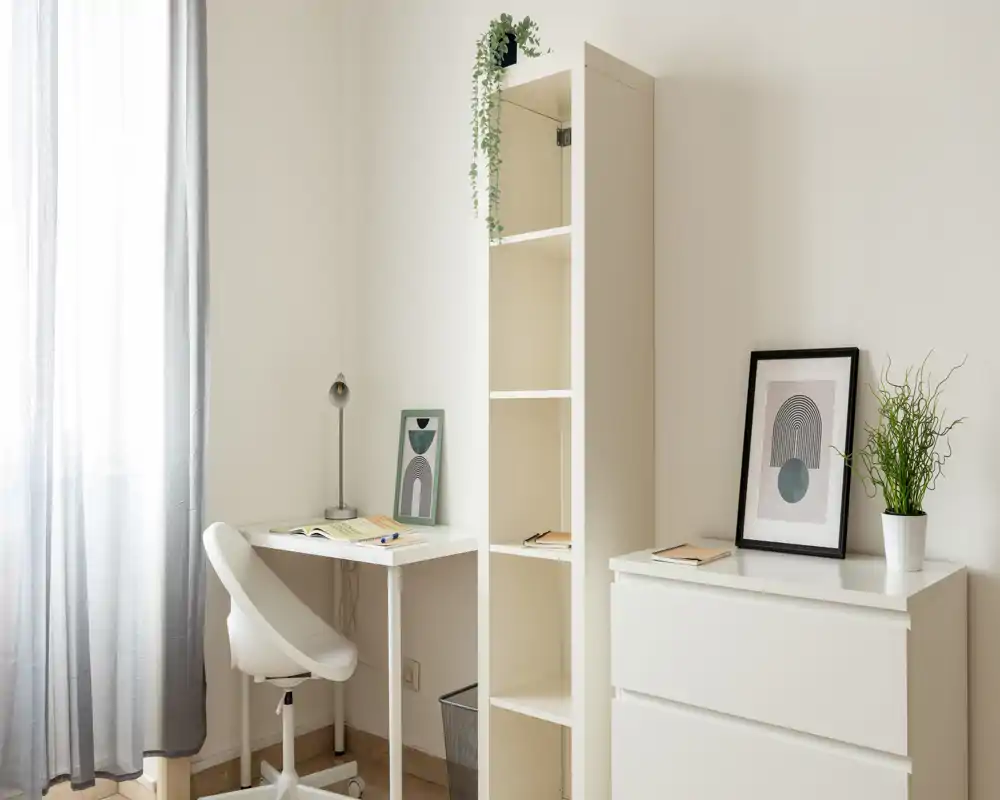 Rincón de trabajo luminoso y minimalista con escritorio blanco, silla ergonómica, estantería alta y detalles decorativos. La luz natural y las cortinas claras hacen el espacio agradable para estudiar o trabajar desde casa.
