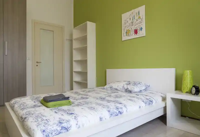 Camera da letto luminosa e ben curata con struttura letto bianca, biancheria a fantasia e parete verde a contrasto. Completano l'ambiente un comodino con lampada, mensole aperte e armadio a muro, perfetta per annunci.