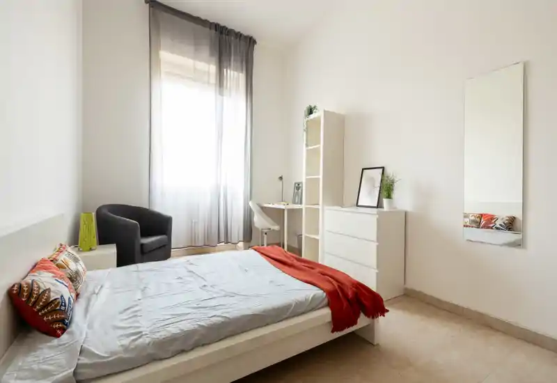 Camera da letto luminosa e minimal con letto matrimoniale, cuscini decorativi e un plaid rosso, poltrona, scrivania e comò vicino a una finestra ampia che illumina la stanza.