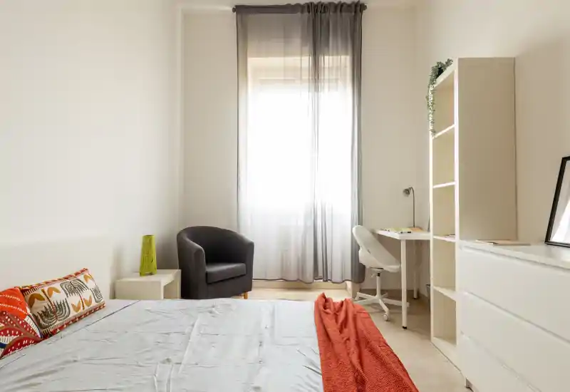 Camera da letto luminosa e minimal con letto confortevole, cuscini decorativi e un caldo plaid arancione, una poltrona vicino alla finestra e una scrivania compatta.