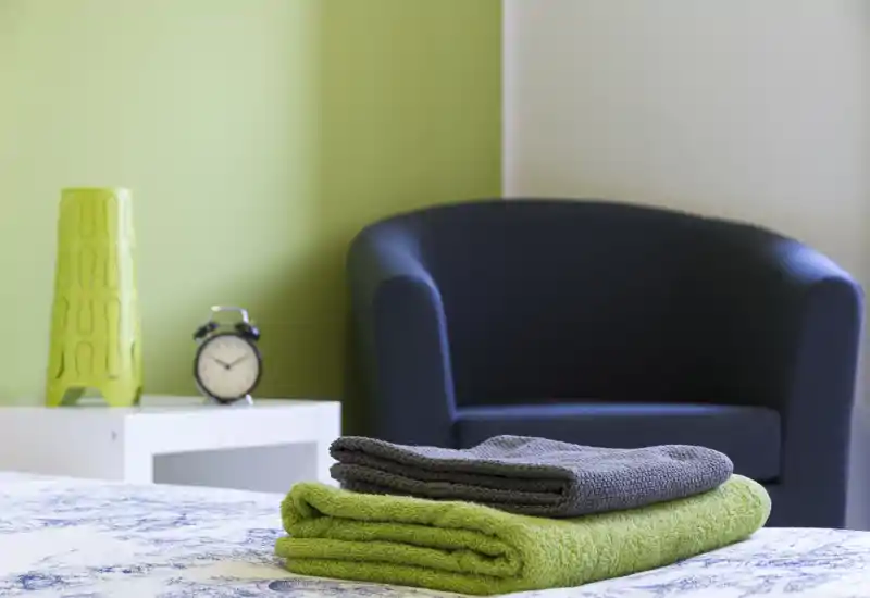 Dettaglio luminoso di una camera da letto moderna con asciugamani piegati sul letto, un comodino con una sveglia e una comoda poltrona sullo sfondo.