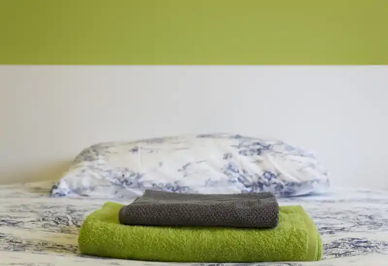 Camera da letto luminosa e ordinata con cuscino a fantasia, biancheria bianco-blu e un mucchio di asciugamani piegati in primo piano; stile pulito e moderno con parete accentuata verde.