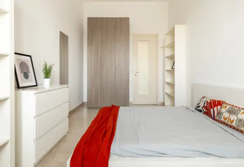 Camera da letto luminosa e minimal con letto matrimoniale, armadio e comò; una coperta rossa e cuscini decorativi aggiungono carattere.