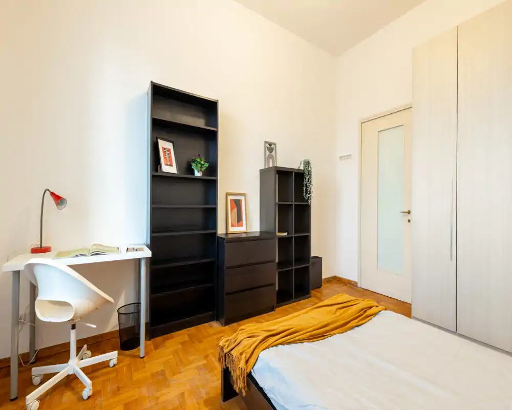 Dormitorio luminoso y ordenado con cama baja moderna, manta mostaza y estanterías sencillas; un pequeño escritorio ofrece espacio de trabajo.