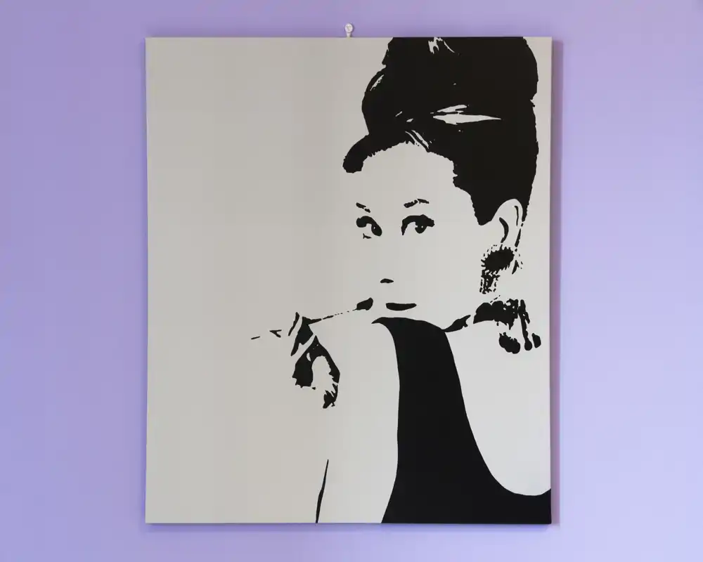 Primo piano di una stampa ritratto in bianco e nero appesa a una parete color lilla. L'opera domina l'inquadratura e non sono visibili mobili o elementi che definiscono la stanza.