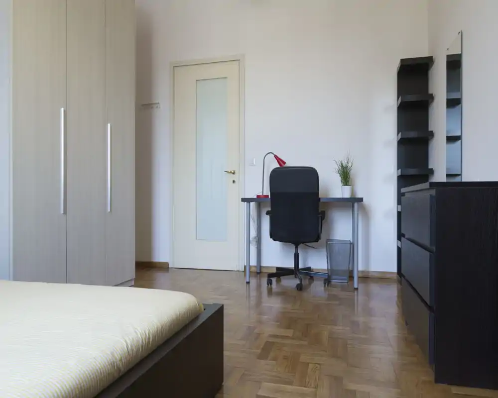 Camera da letto luminosa e minimale con letto ben visibile in primo piano, armadio, scrivania e scaffali — pavimento in parquet e arredo neutro e ordinato.