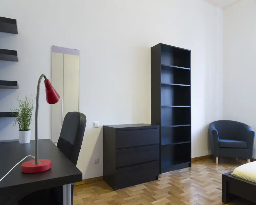 Camera da letto luminosa e minimale con mobili neri moderni: comò, libreria alta, scrivania con lampada rossa e angolo del letto visibile. Pavimento in parquet e pareti neutre creano uno spazio funzionale e tranquillo, ideale per affitti brevi.