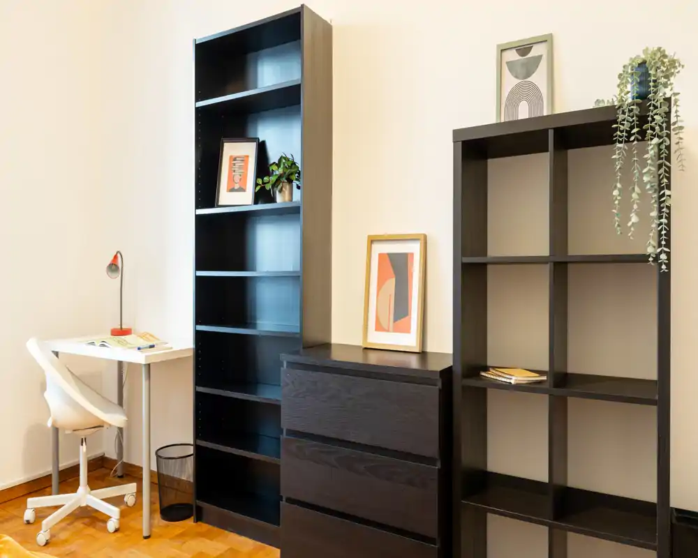 Rincón de dormitorio luminoso con estanterías oscuras, cómoda y un pequeño escritorio blanco con silla; láminas decorativas y una planta aportan un estilo moderno y ordenado.